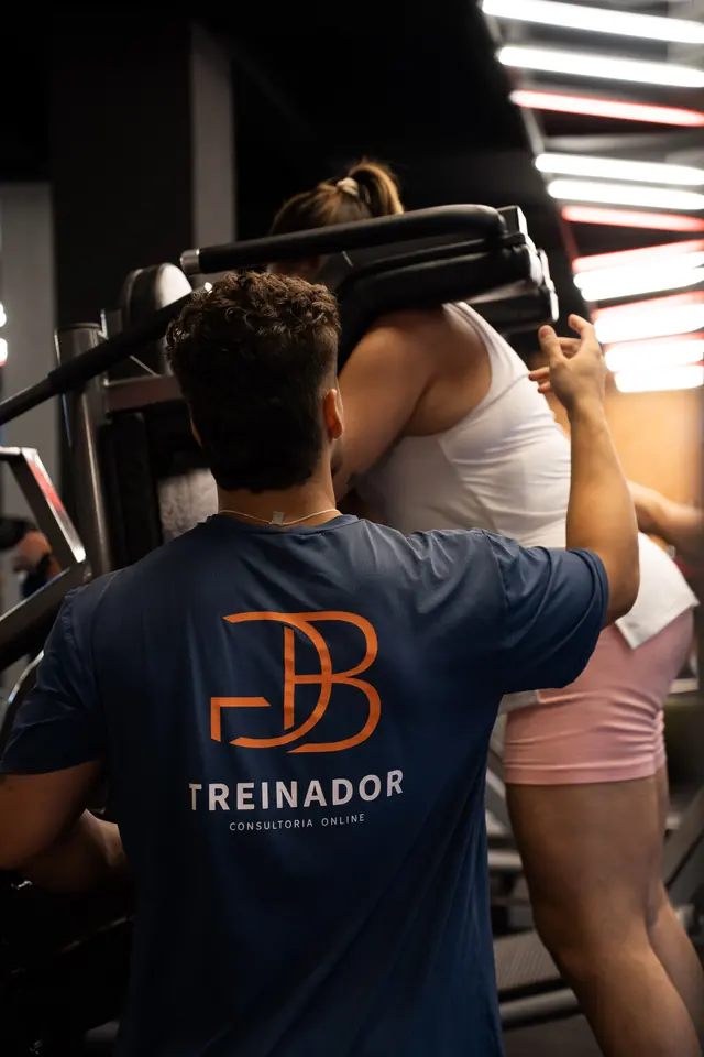 Gustavo Borba de costas com a camiseta Treinador Consultoria Online orientando uma aluna na máquina